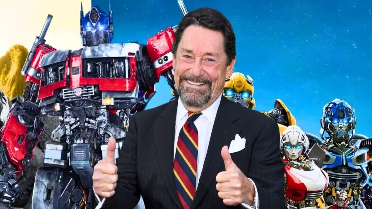 Peter Cullen in a dark suit and striped tie, smiling and giving two thumbs up, composited in front of Optimus Prime, Bumblebee, Arcee and Mirage.