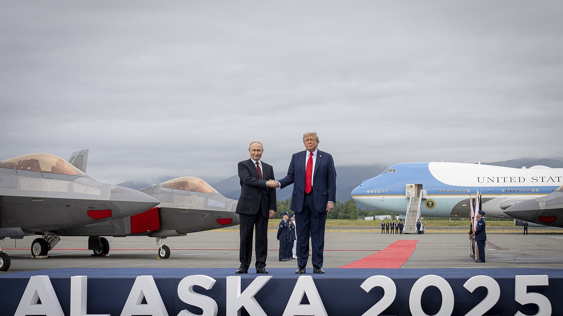 Donald Trump and Vladimir Putin shake hands on a stage reading ALASKA 2025, with F-22 fighter jets on the left and Air Force One behind them.