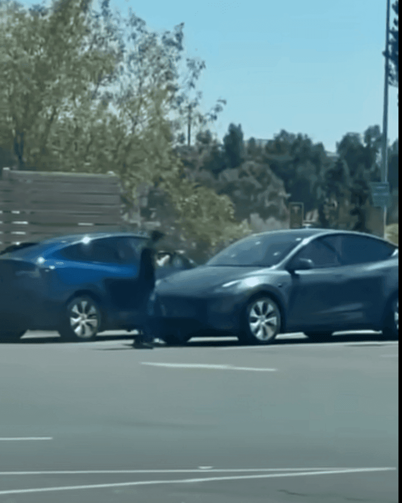 A distant, low-resolution frame showing a person standing beside a blue car in a car park, with a second dark car in the foreground.