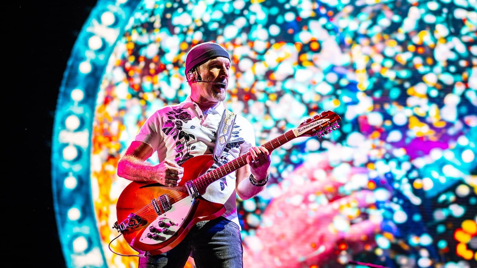 U2 guitarist The Edge in a beanie and floral shirt plays a red Rickenbacker guitar in front of a huge circular screen of colourful dots.