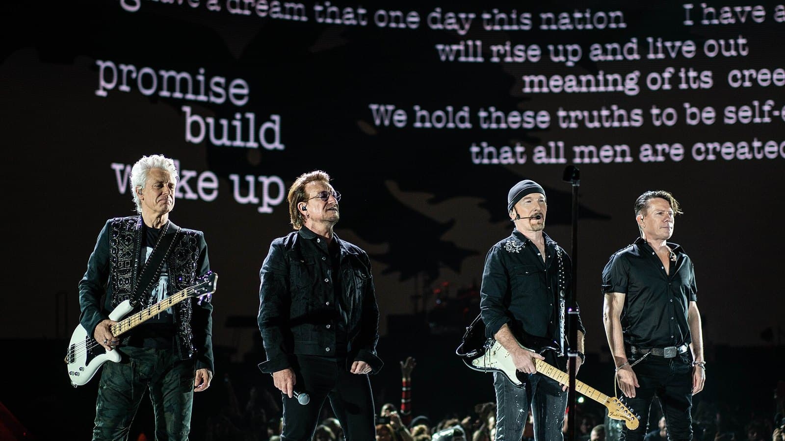 The four members of U2 stand in a line at the front of a stadium stage in black clothes, with lines of text projected on a giant screen behind them.