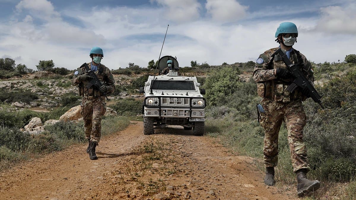 Two Italian soldiers wearing blue UN helmets and face masks walk along a dirt track in southern Lebanon, followed by a white armoured vehicle.