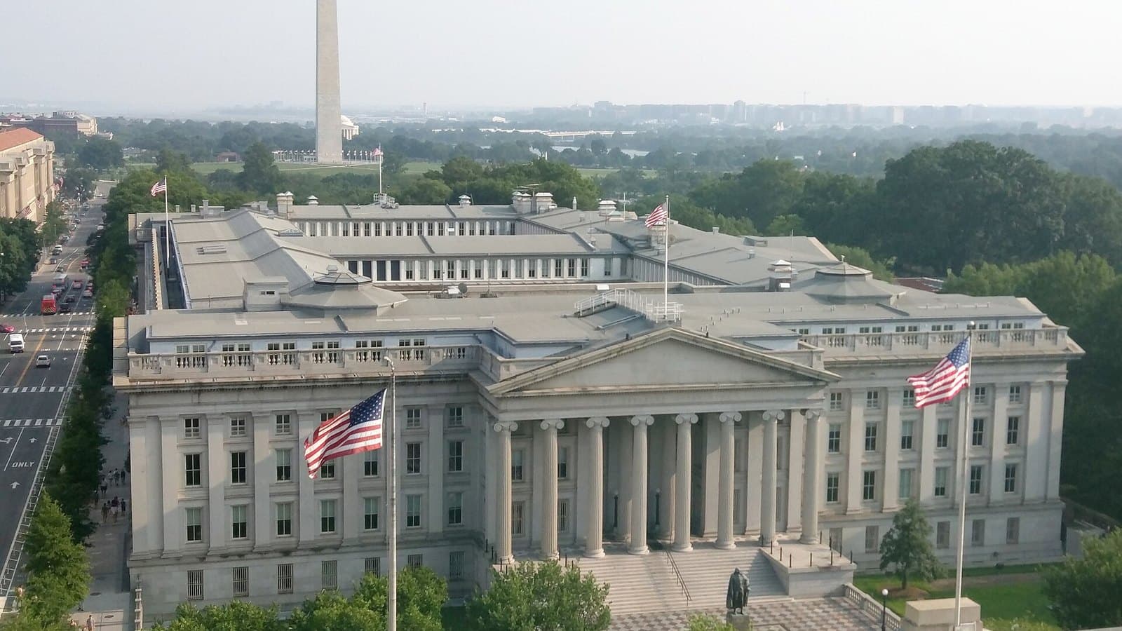Elevated view of the grey stone U.S. Treasury Building with columns and two American flags, surrounded by trees, with the Washington Monument rising in the hazy background.