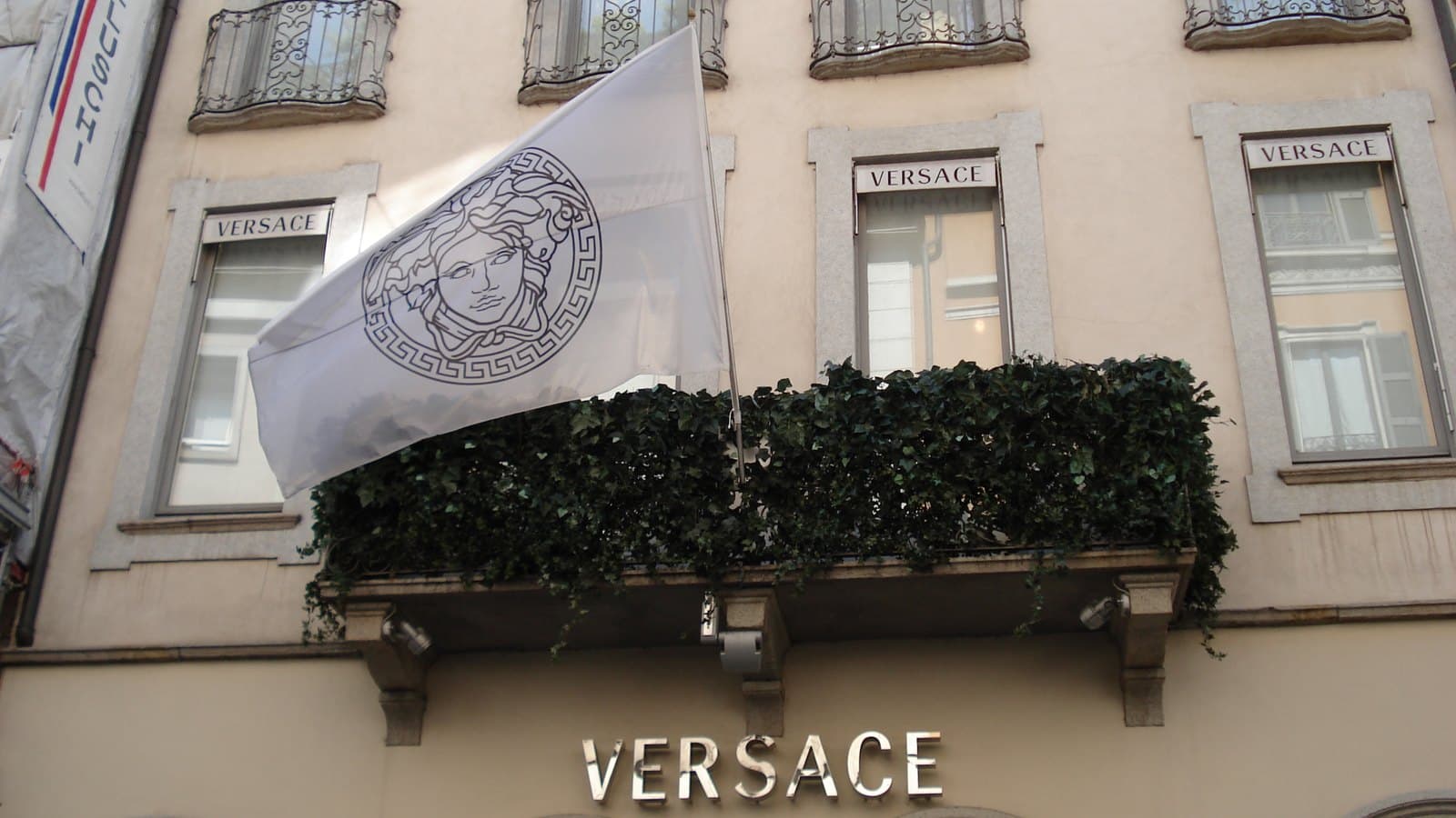 The cream stone facade of the Versace boutique on Via Montenapoleone in Milan, with a white Medusa-logo flag, a hedge-lined balcony and Versace lettering above the entrance.