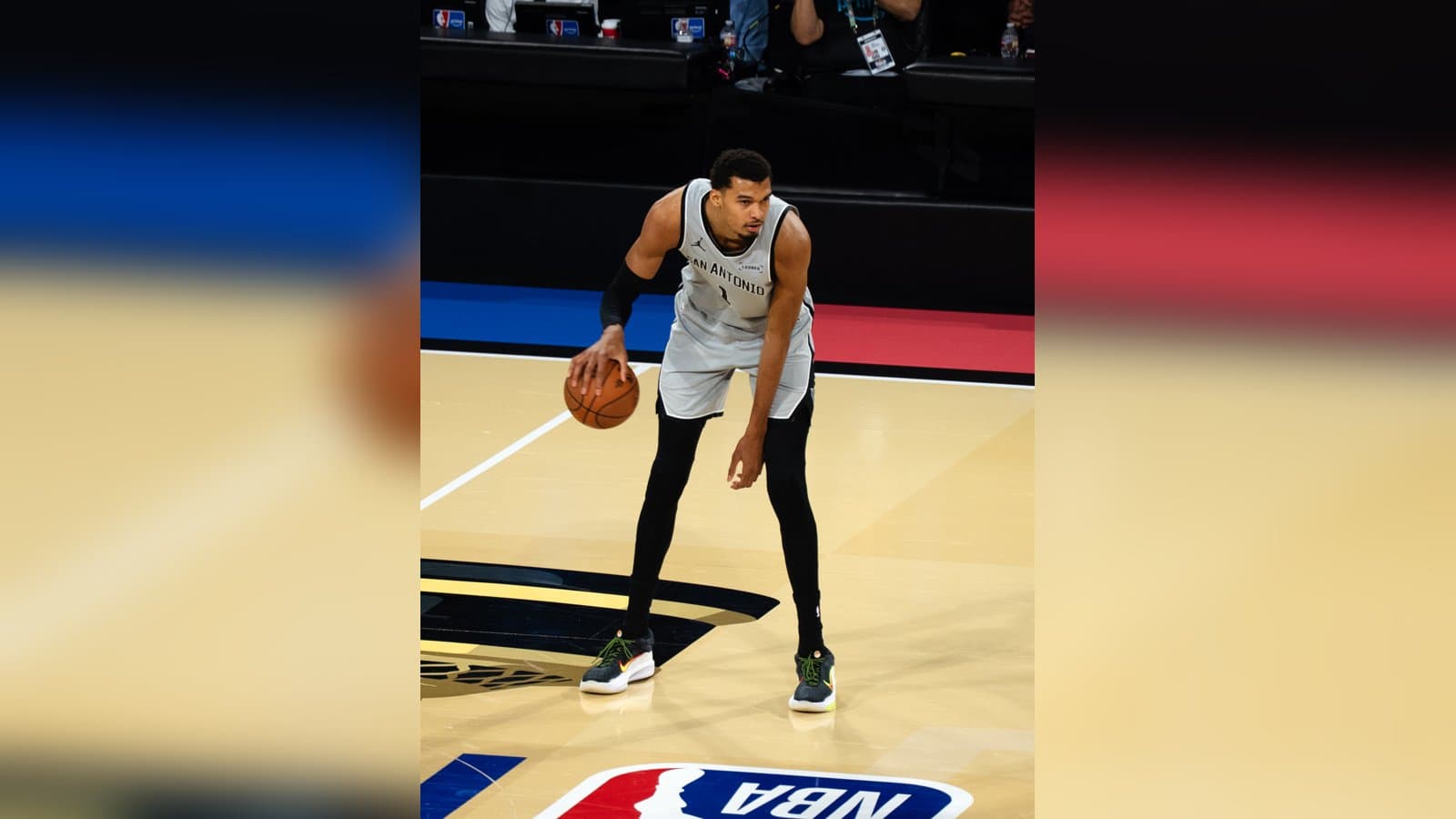 Victor Wembanyama in a white Spurs uniform dribbling the basketball near the NBA logo on the court.