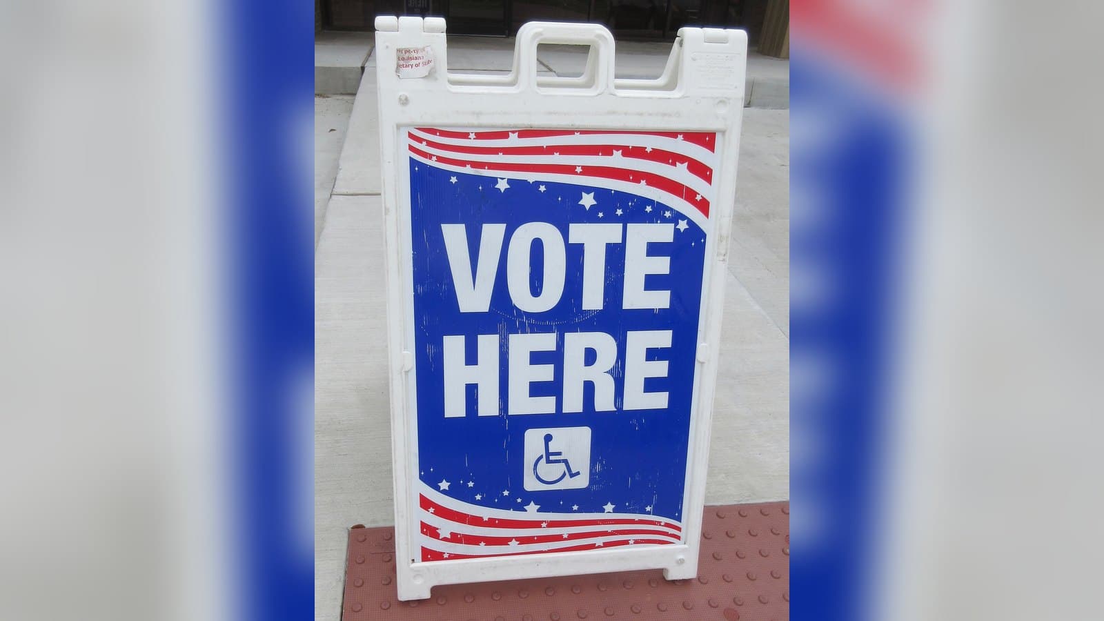 A white sandwich-board sign reading VOTE HERE in large blue letters with red stripes and a wheelchair-access symbol, standing on a sidewalk outside a polling place.