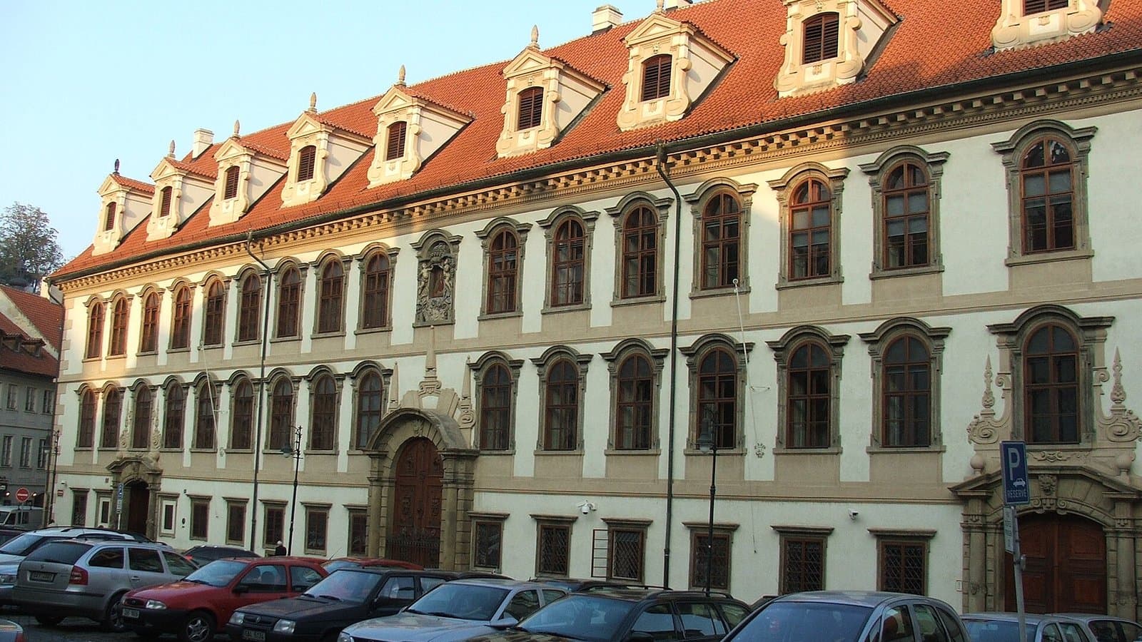 The pale yellow baroque facade of Wallenstein Palace in Prague, with a red-tiled roof and dormer windows, and cars parked in the square in front.