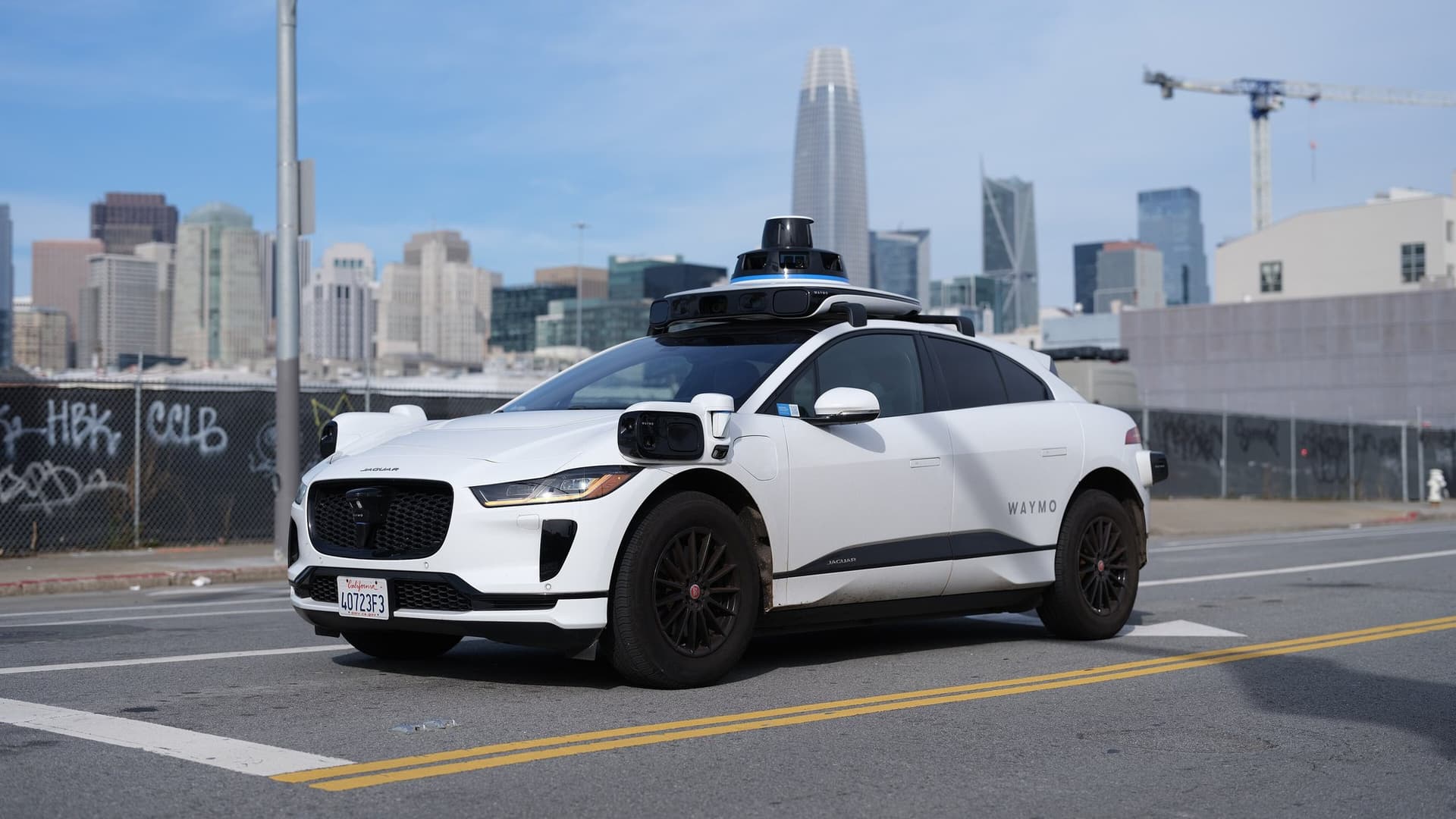 A white Waymo Jaguar I-Pace robotaxi with a roof-mounted sensor dome on a San Francisco street, with the city skyline and Salesforce Tower behind it.