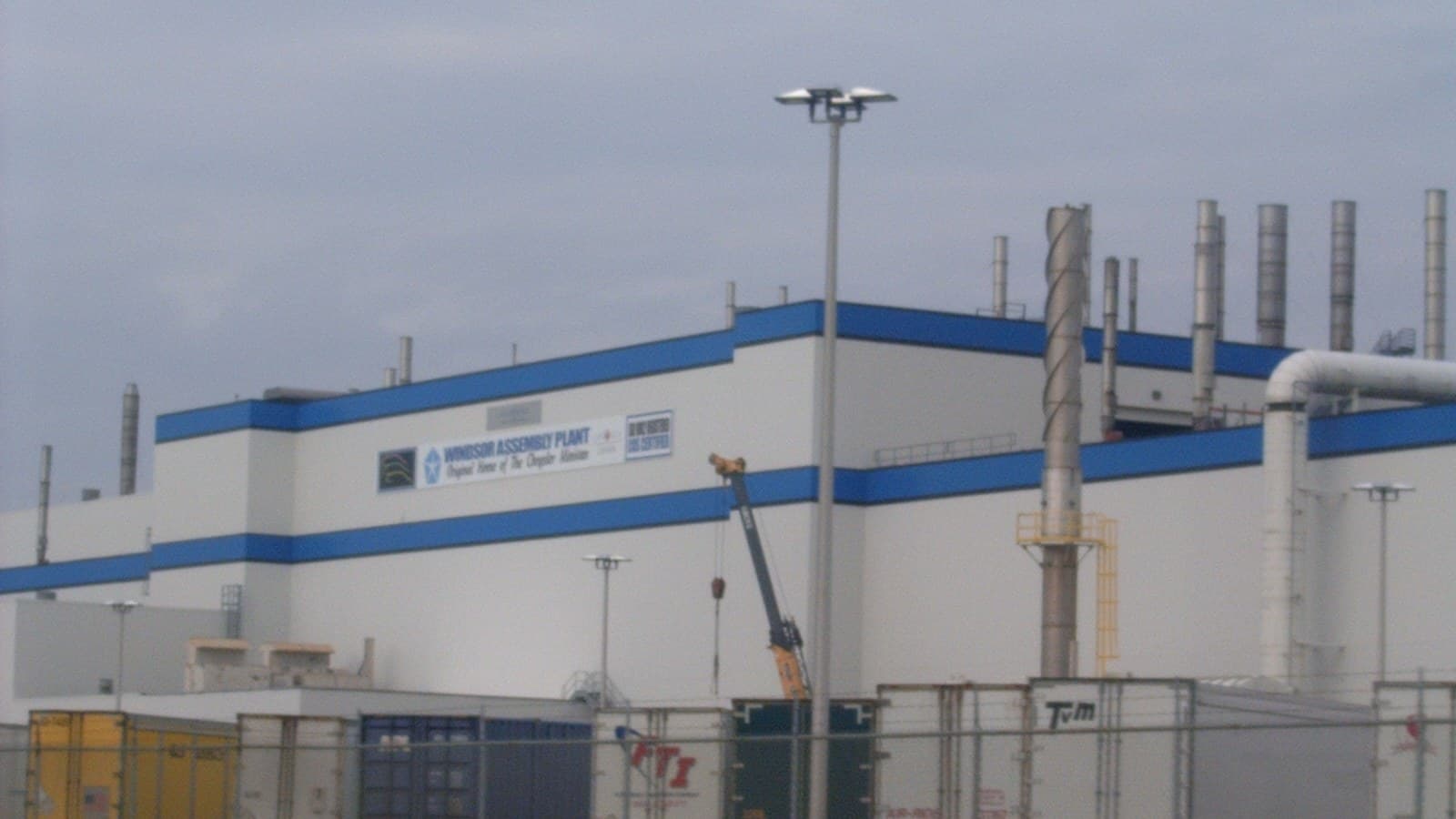 Exterior of the white and blue Windsor Assembly Plant building in Windsor, Ontario, with its sign, exhaust stacks and parked truck trailers in front.