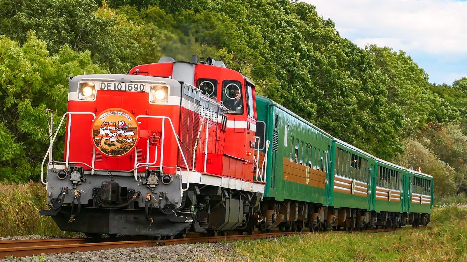 A red DE10 diesel locomotive pulling green and brown observation coaches of the Norokko sightseeing train through summer greenery in eastern Hokkaido.