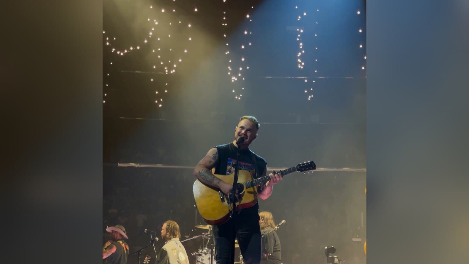 Zach Bryan sings and plays acoustic guitar on an arena stage under warm lights.