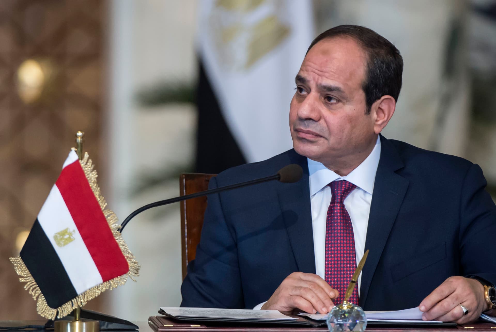 Abdel Fattah al-Sisi in a dark suit speaking at a desk with an Egyptian flag beside him.