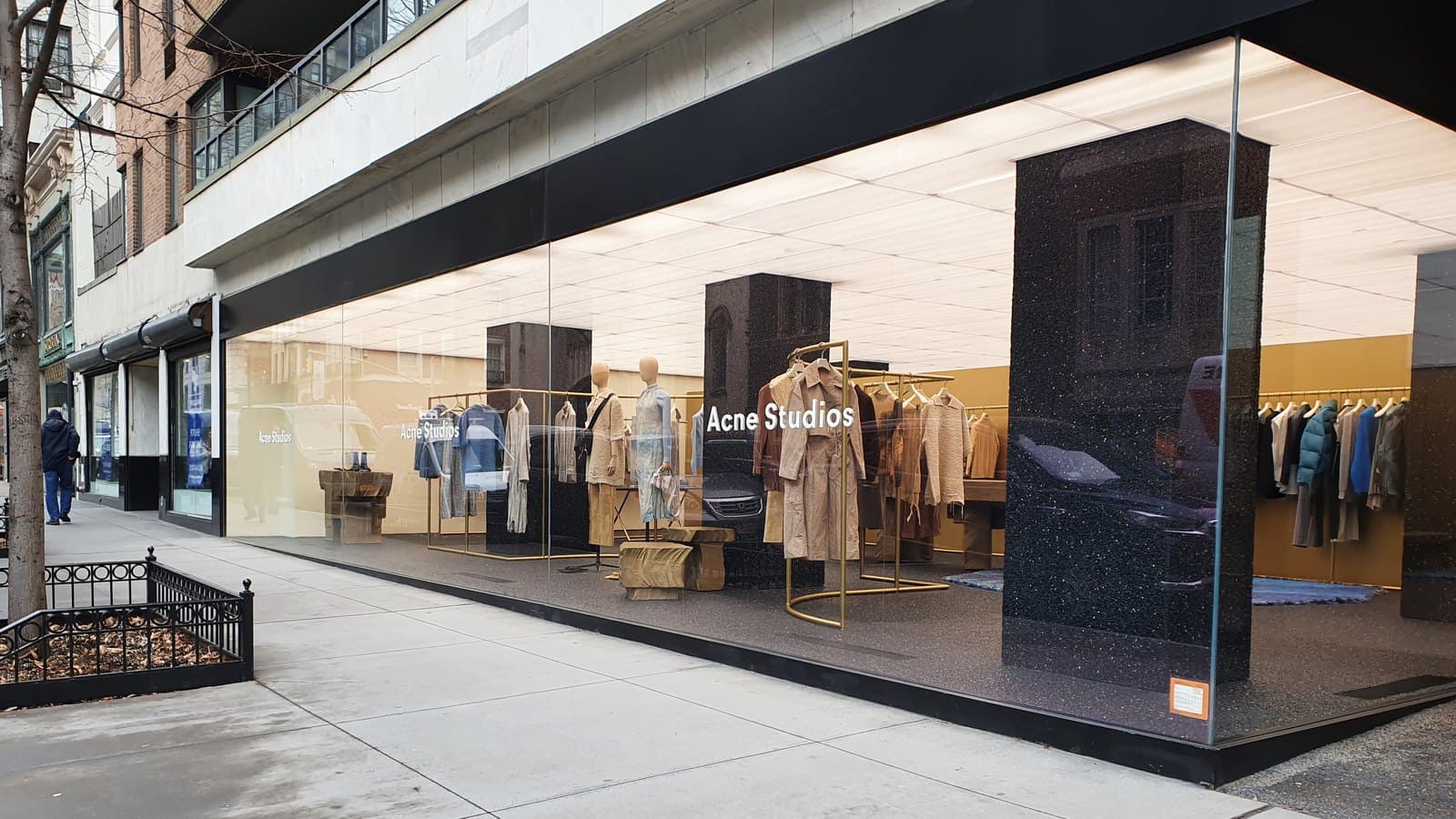 A corner Acne Studios boutique with floor-to-ceiling glass windows, showing racks of coats and knitwear and a mannequin display on a city sidewalk.