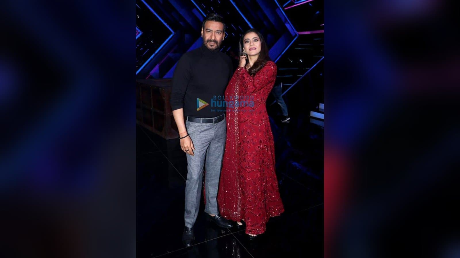 Ajay Devgn, in a black turtleneck and grey trousers, poses beside Kajol, in a red embroidered outfit, on a television studio set.