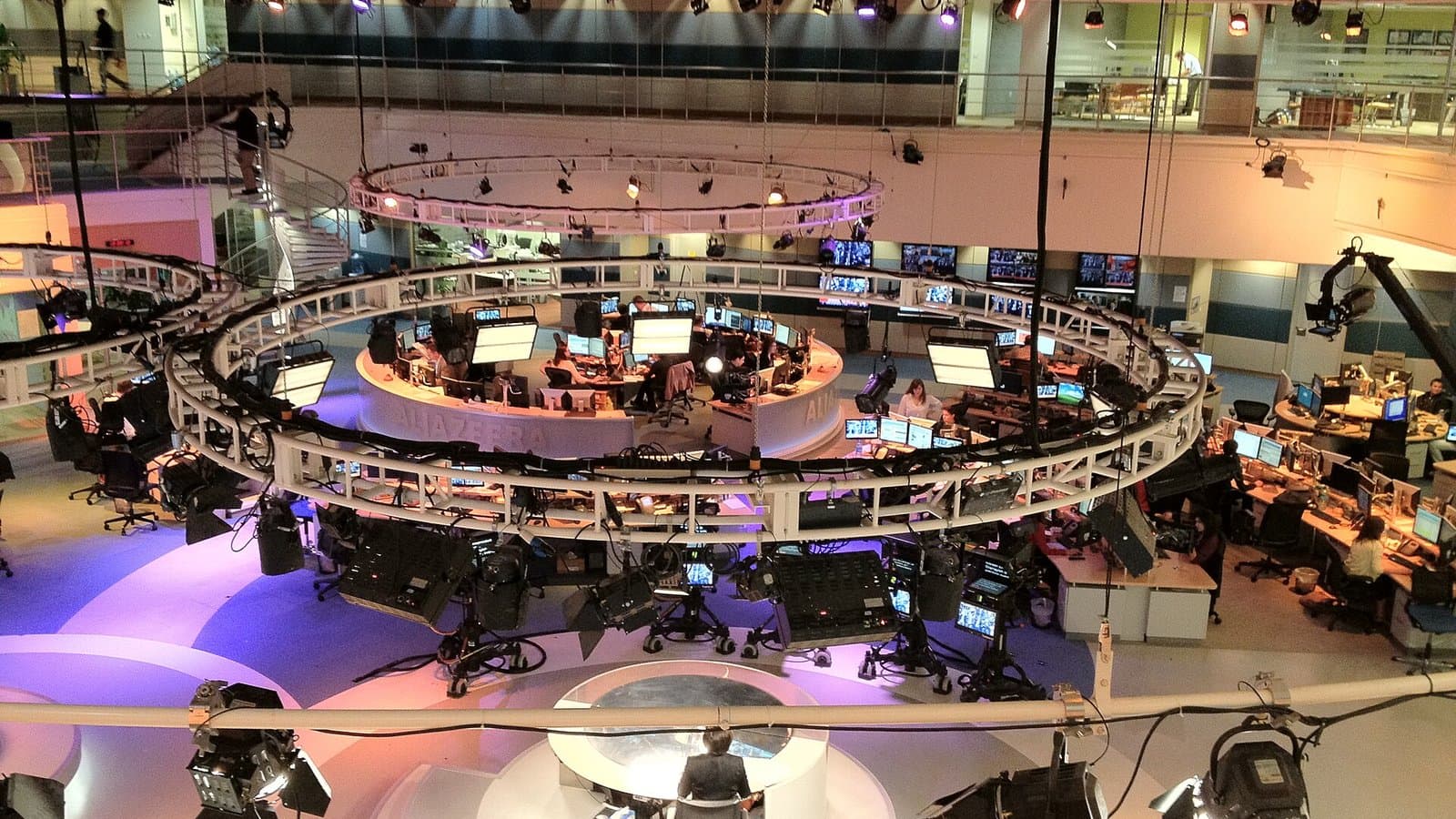 Overhead view of the circular Al Jazeera English newsroom in Doha, with rows of journalists' desks and screens around a central studio.