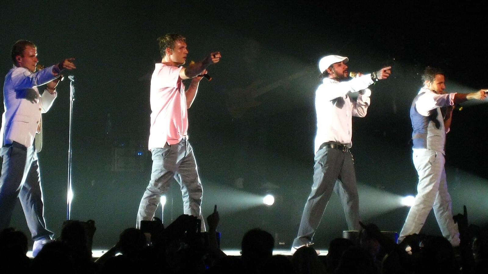 Four members of the Backstreet Boys in light-coloured shirts point toward the crowd from the front of a concert stage, lit by white spotlights.