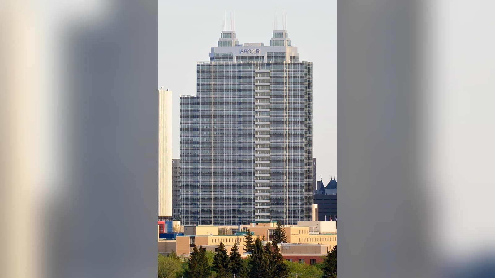 Epcor Tower, a tall glass office skyscraper with twin rooftop spires and an EPCOR sign, rising above trees and low buildings in Edmonton