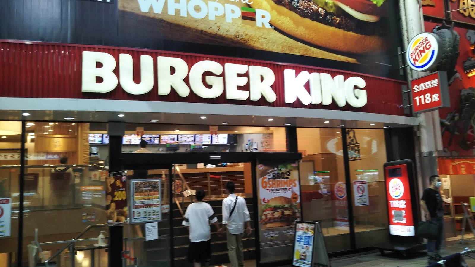The red-and-white storefront of a Burger King restaurant in Namba, Osaka, with a Whopper billboard above the entrance and customers walking in.