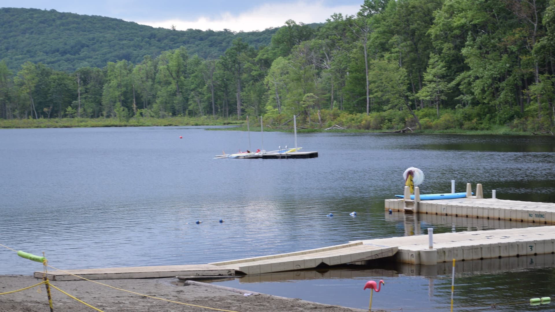 A calm lake ringed by thick green forest, with a floating dock, a moored raft of small sailboats and a sandy beach in the foreground at Camp No-Be-Bo-Sco in New Jersey.