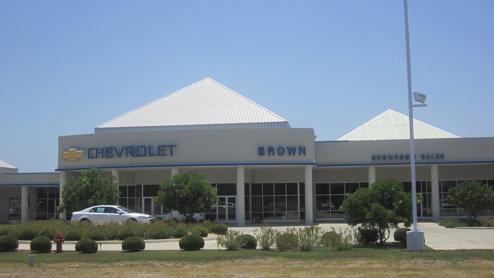 A small-town Chevrolet dealership building with new pickup trucks and cars parked out front under a blue sky.