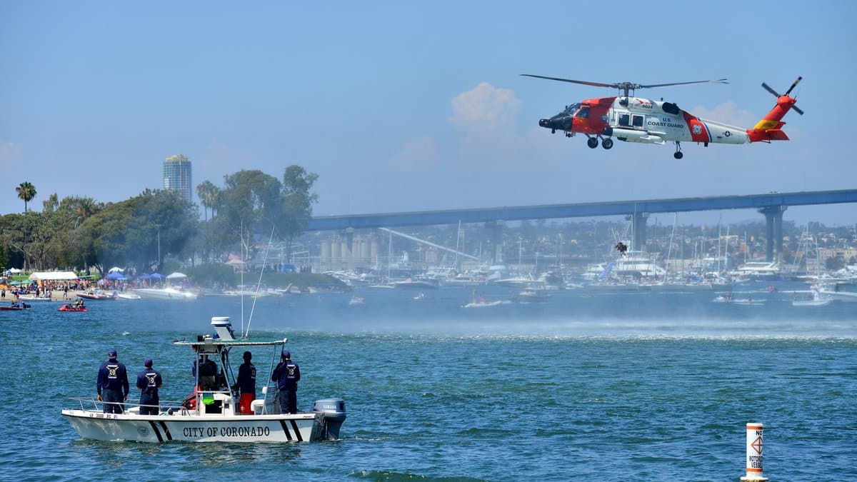An orange and white U.S. Coast Guard MH-60 Jayhawk helicopter hovering low over water during a rescue demonstration.