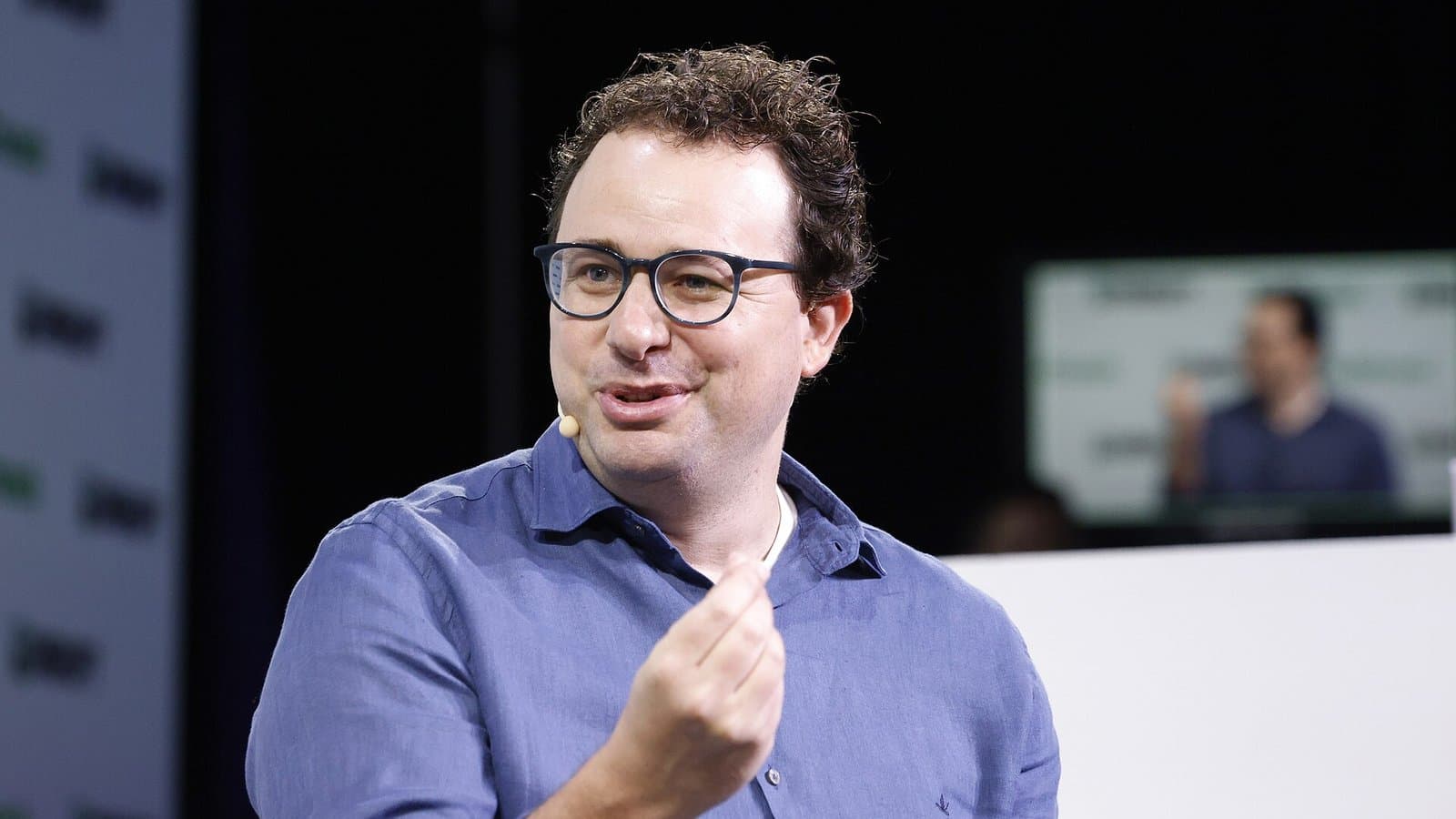 Anthropic CEO Dario Amodei, wearing glasses and a blue shirt, speaking and gesturing on stage at TechCrunch Disrupt 2023.