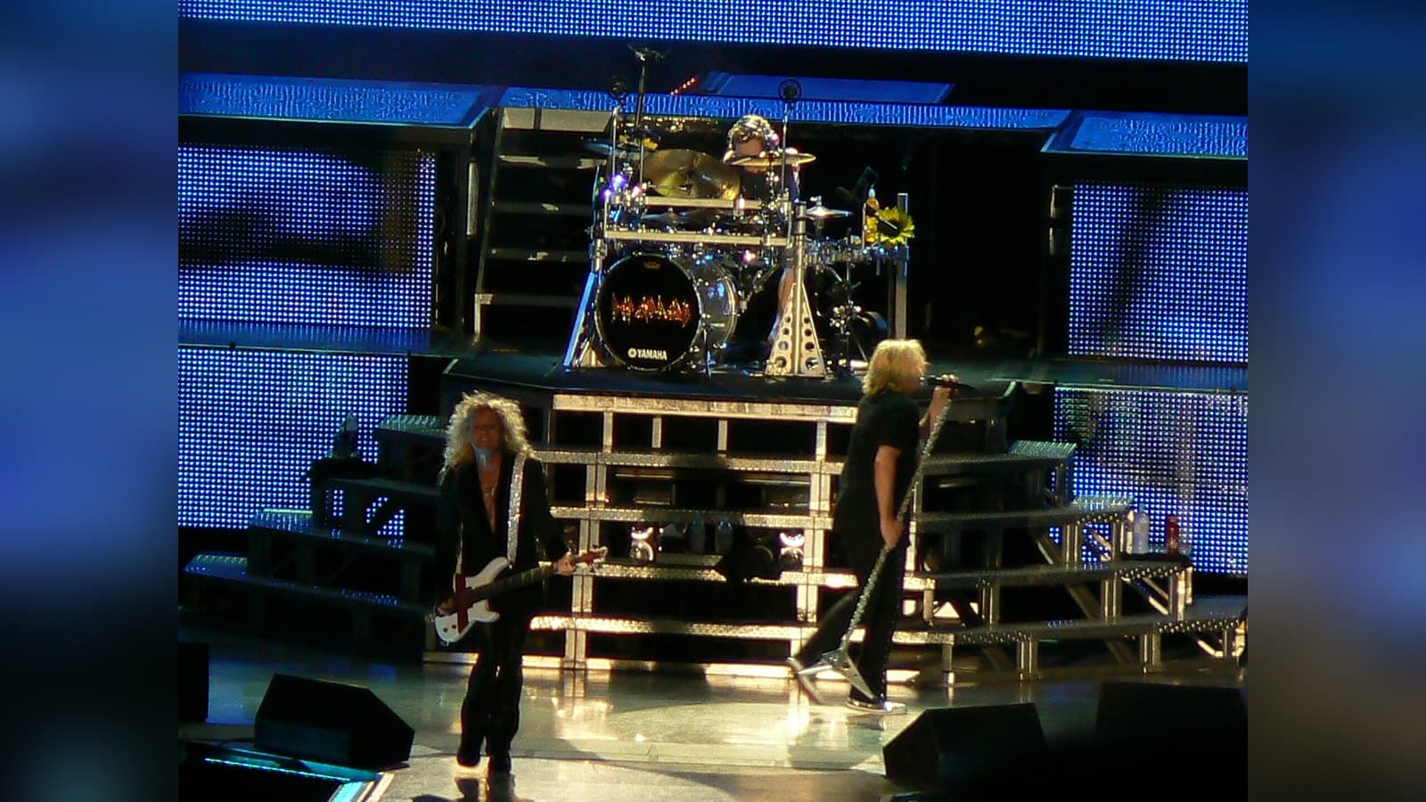 Def Leppard's bassist and singer perform on a tiered silver stage in front of a drum riser and a glowing blue LED backdrop.
