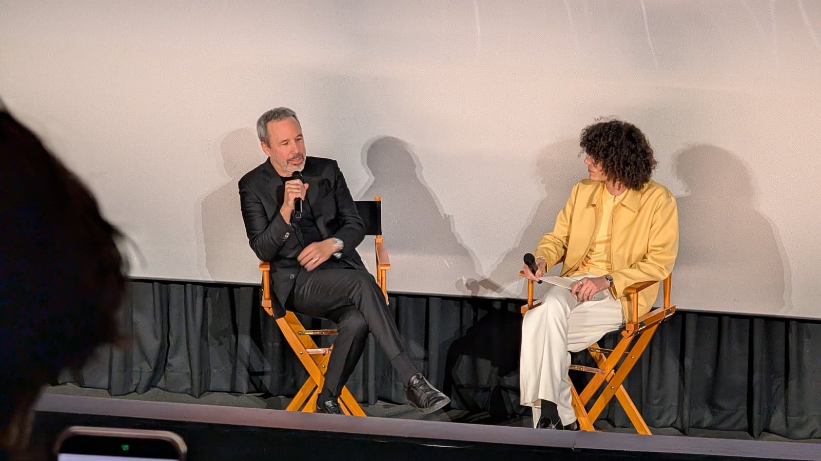 Director Denis Villeneuve, in a black suit, speaks into a microphone from a director's chair on a cinema stage beside a moderator in a yellow jacket.