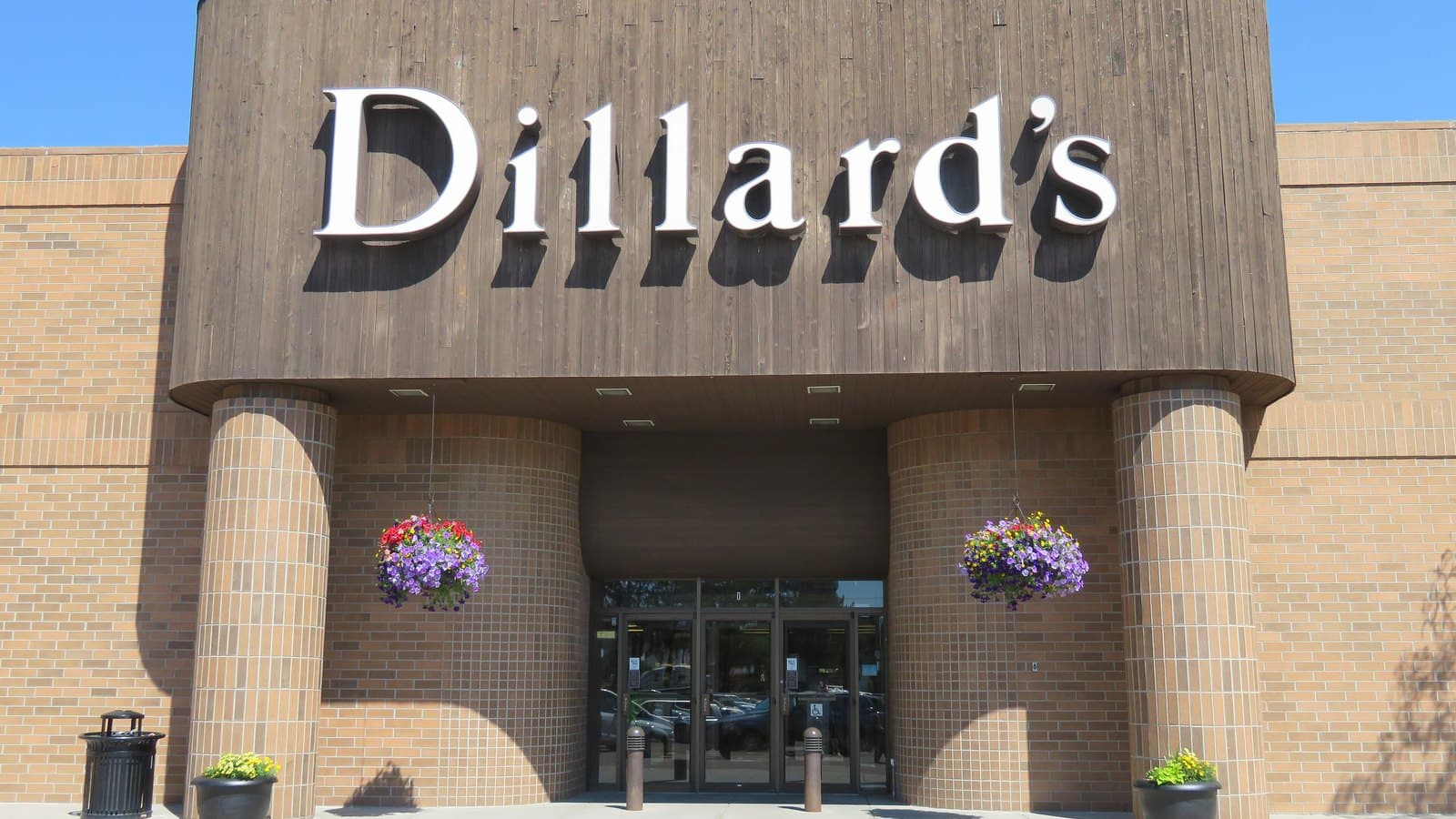 The entrance of a Dillard's department store with the brand name in white letters on a brown facade, brick columns and hanging flower baskets.