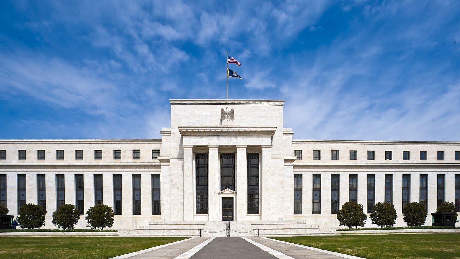 The white marble Eccles Building of the Federal Reserve in Washington under a bright blue sky with wispy clouds, an American flag flying above its columned central entrance and a row of trimmed shrubs in front.