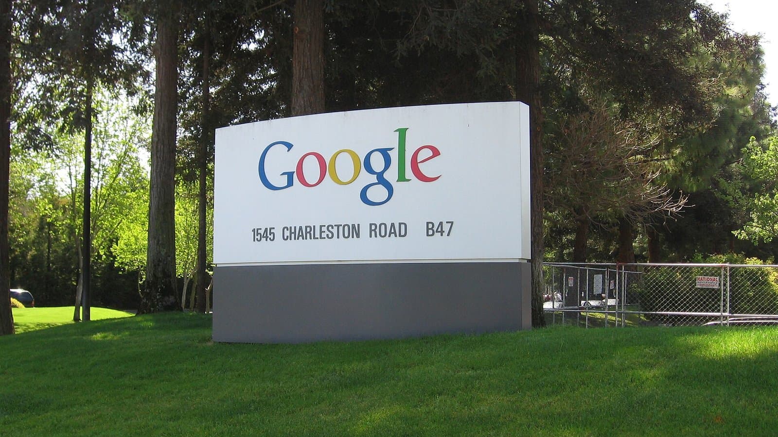 A white Google sign with the colorful Google logo and the address 1545 Charleston Road on a lawn in front of trees at the Googleplex.