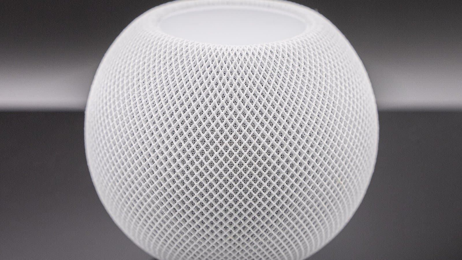 Close-up of a round white Apple HomePod mini smart speaker with its mesh fabric cover against a dark gray background.
