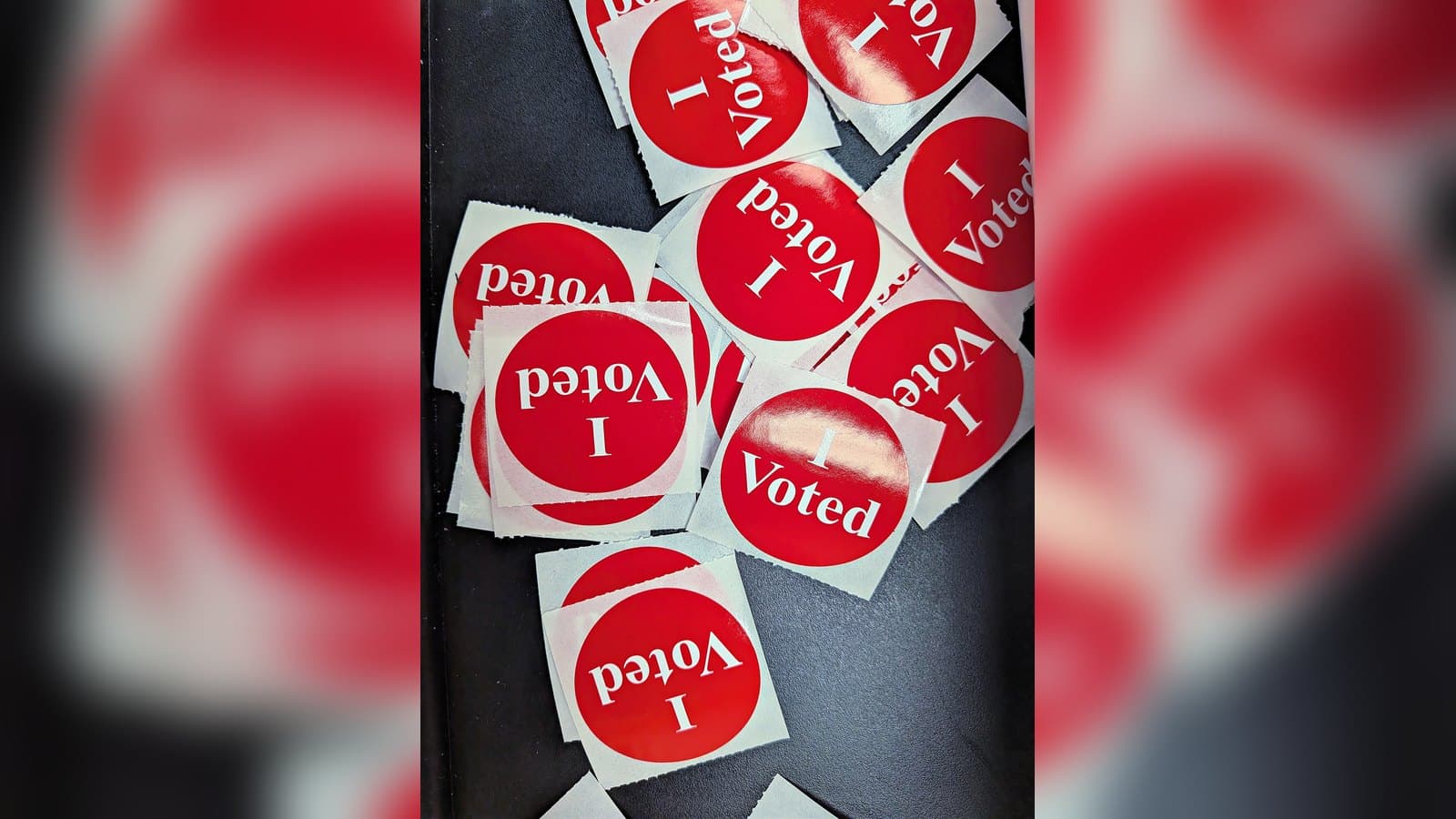 A scattered pile of round red and white I Voted stickers on a dark surface at a polling station.