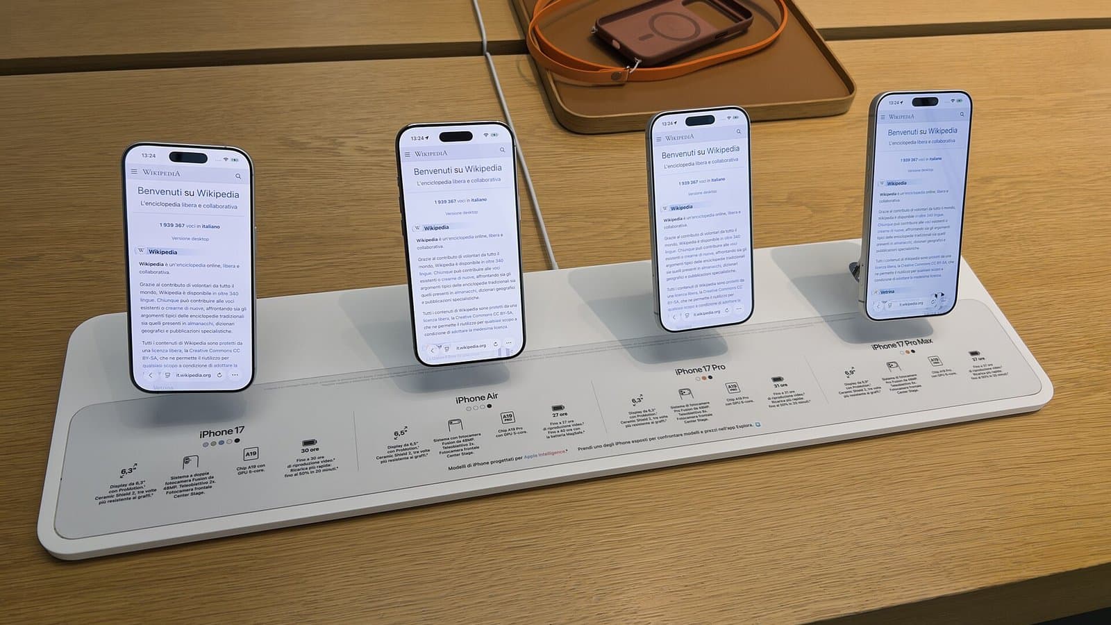 Four iPhones from the iPhone 17 lineup standing upright on a white display stand on a wooden Apple Store table.