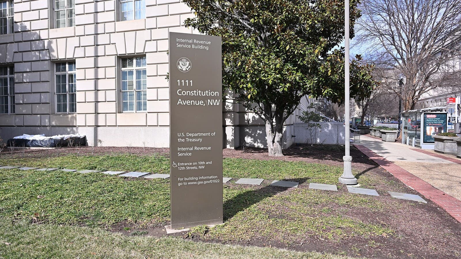 A tall brown sign on a lawn reading Internal Revenue Service Building, 1111 Constitution Avenue NW, U.S. Department of the Treasury, Internal Revenue Service, in front of a pale stone government building and a magnolia tree.