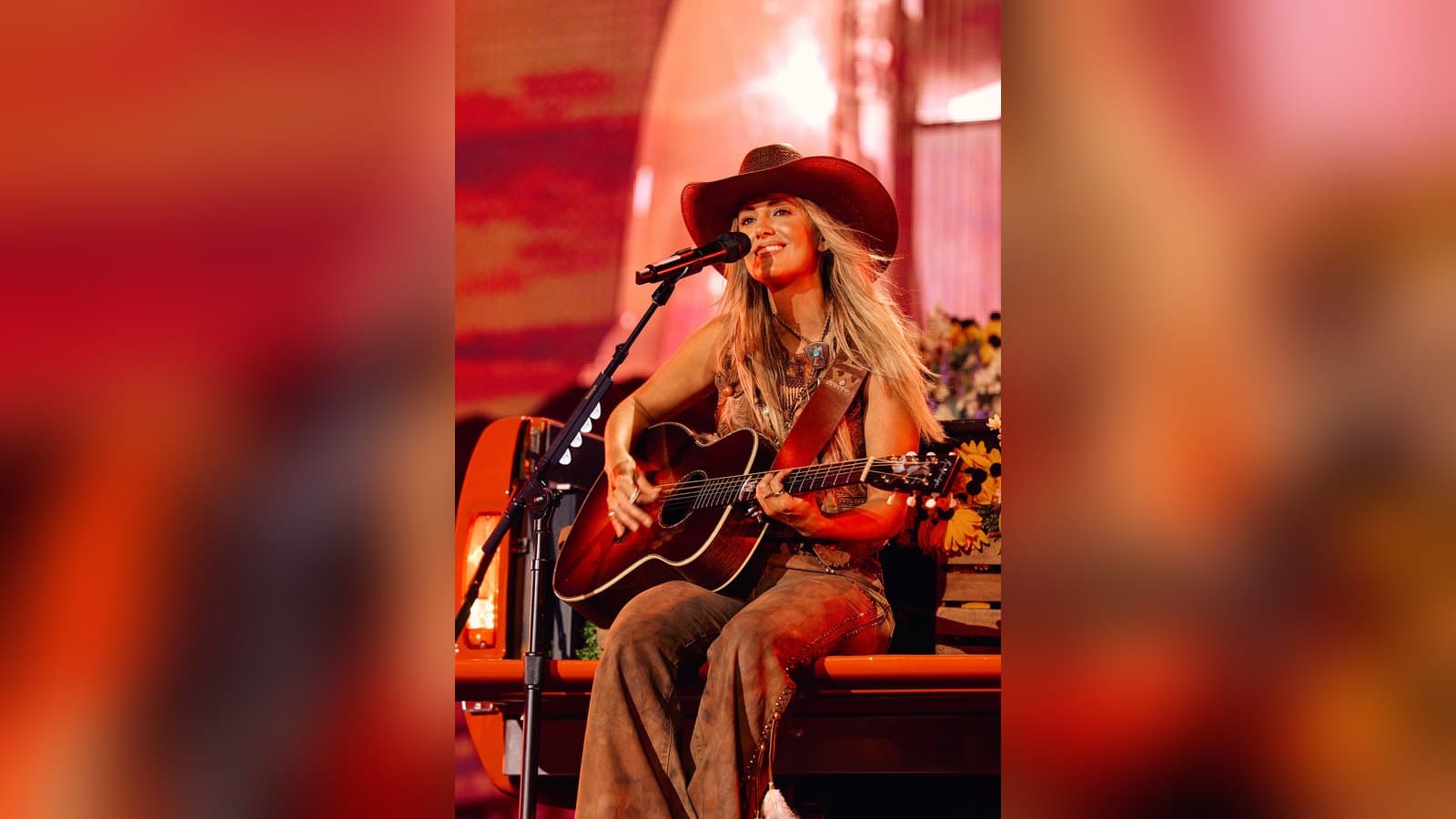Lainey Wilson in a wide-brimmed cowboy hat and flared trousers sits on stage playing an acoustic guitar and singing into a microphone under warm red light.