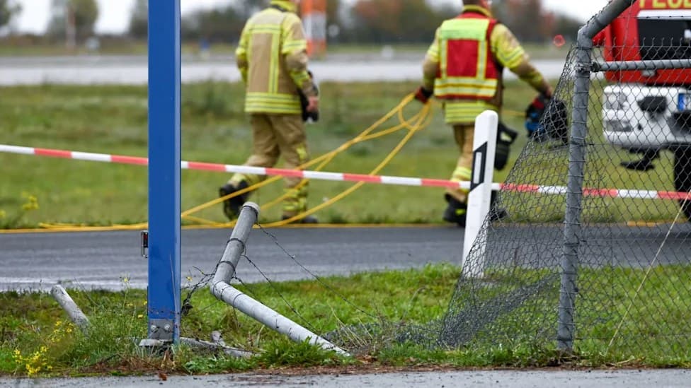 Firefighters and police officers in high-visibility jackets crouch beside a section of flattened wire fence and bent posts at the edge of an airfield.