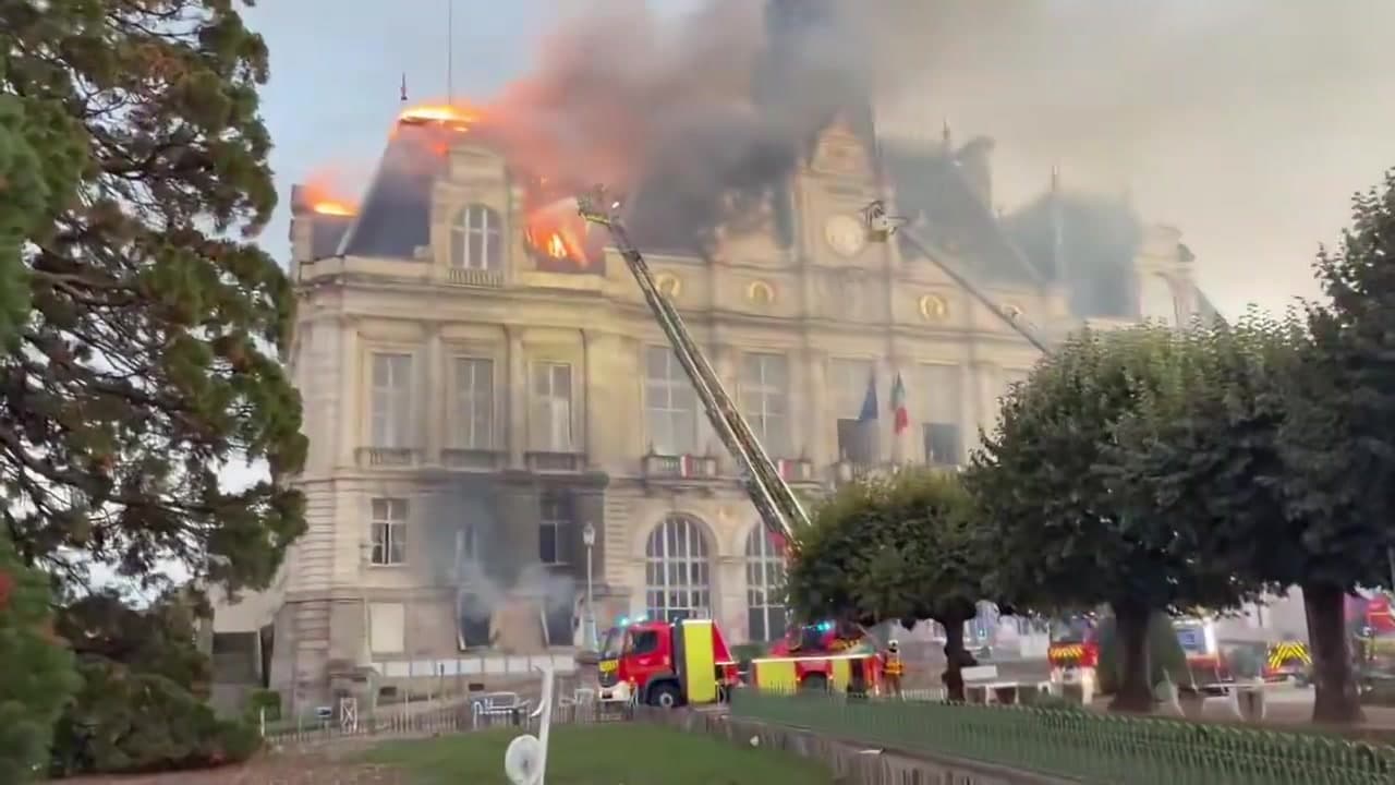 Flames and black smoke pouring from the roof of the ornate stone Limoges city hall as two fire ladders reach toward it and fire trucks wait below.