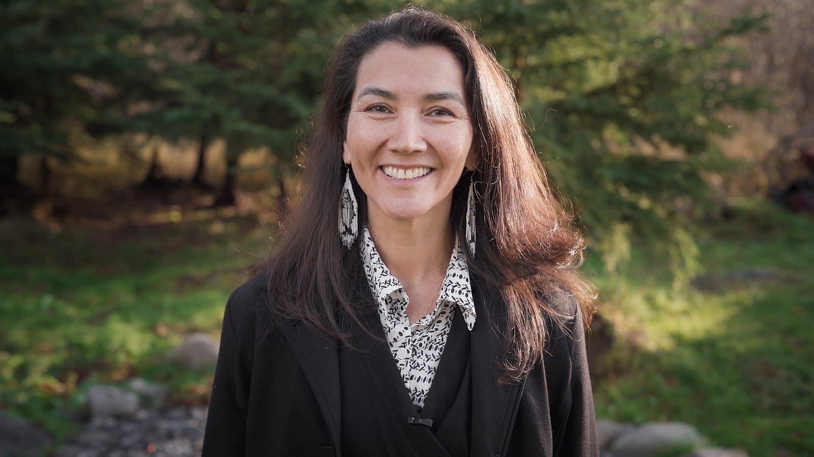 Mary Peltola, smiling, in a dark blazer and patterned blouse, photographed outdoors in front of trees.