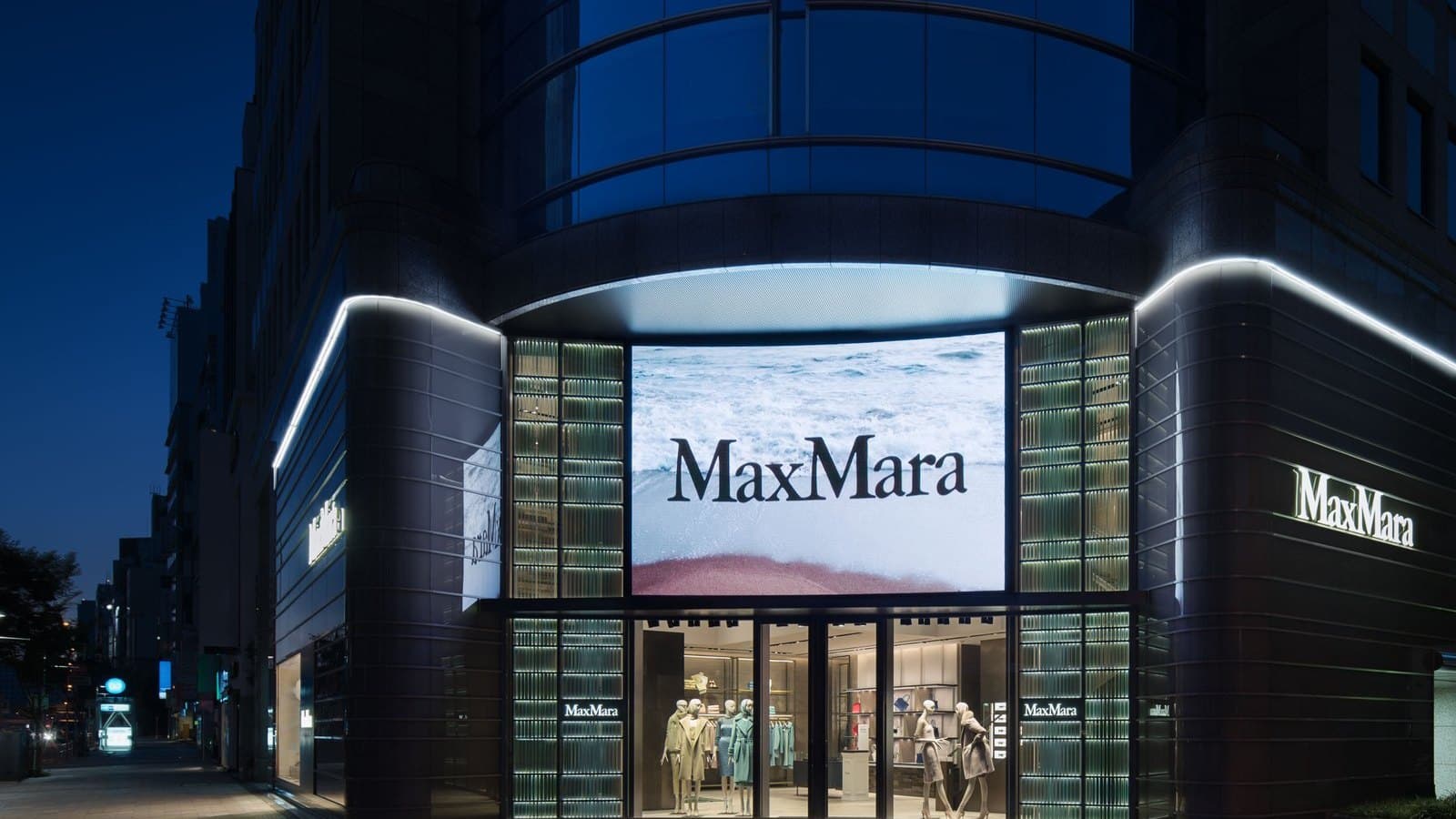 The illuminated glass exterior of a Max Mara boutique in Tokyo's Minami-Aoyama district at dusk, with mannequins dressed in coats in the windows.