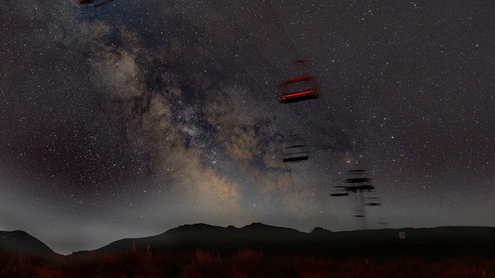 The bright core of the Milky Way rising over dark mountain ridges, with a red-lit ski chairlift in the foreground at night.