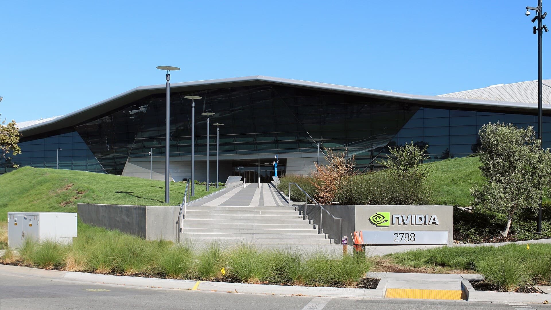 Nvidia's headquarters in Santa Clara, California: a low, angular glass building with a sloping silver roof, concrete steps and a sign reading NVIDIA 2788 under a clear blue sky.