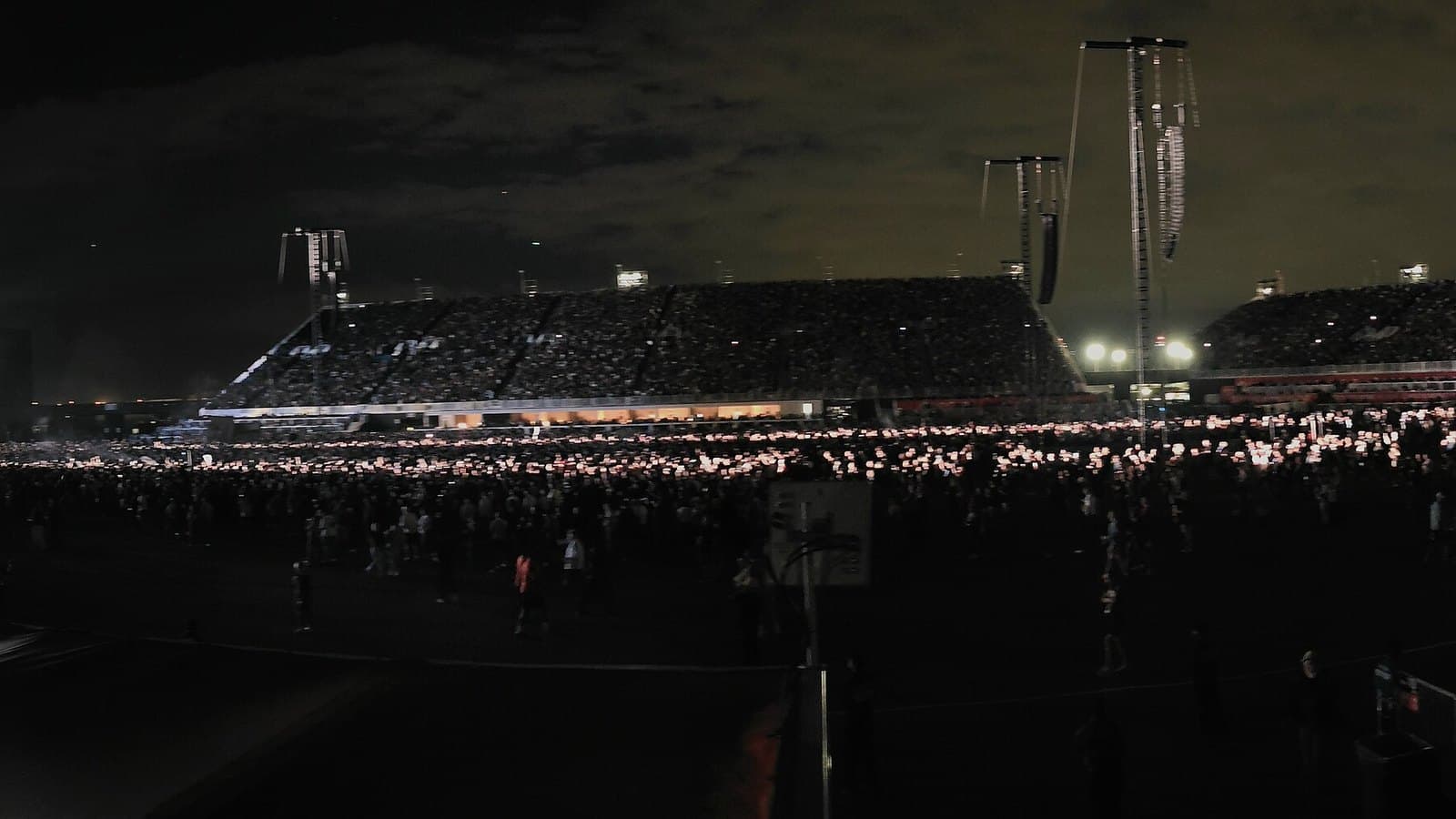 A night-time stadium crowd stretches across a vast open-air field toward a temporary grandstand, lit by thousands of phone lights.