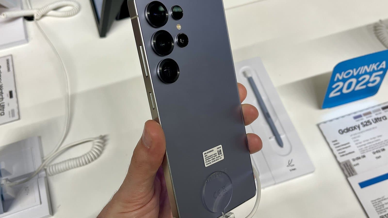 A hand holding a titanium silver-blue Samsung Galaxy S25 Ultra, showing its rear camera lenses, at a store display.