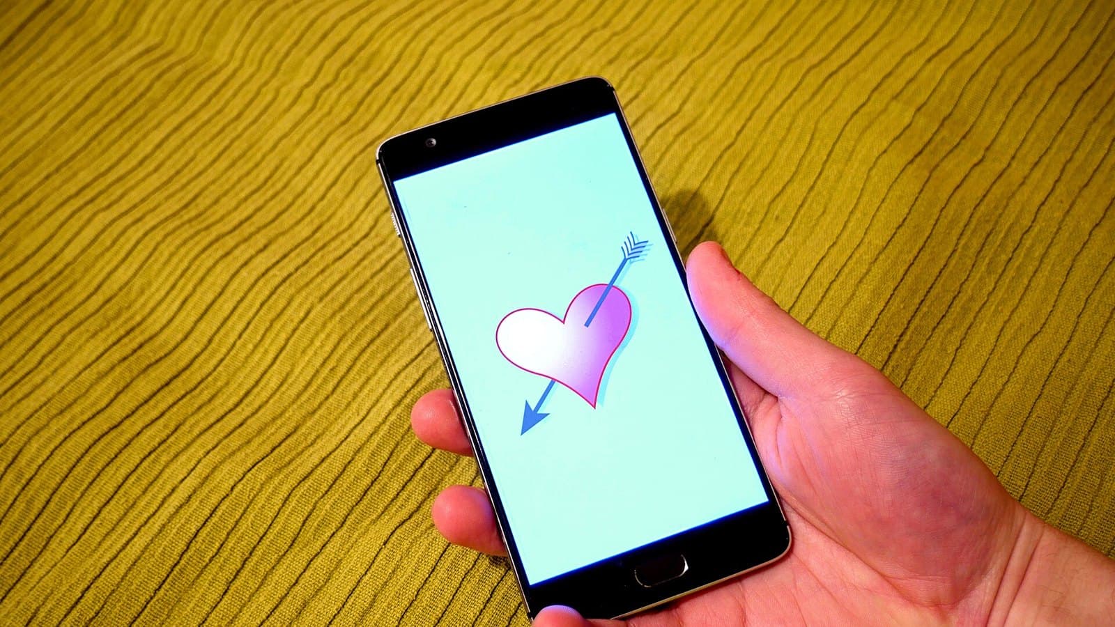 A hand holding a black smartphone against a yellow textured background, the screen showing a pink heart pierced by a blue arrow on a light blue background.