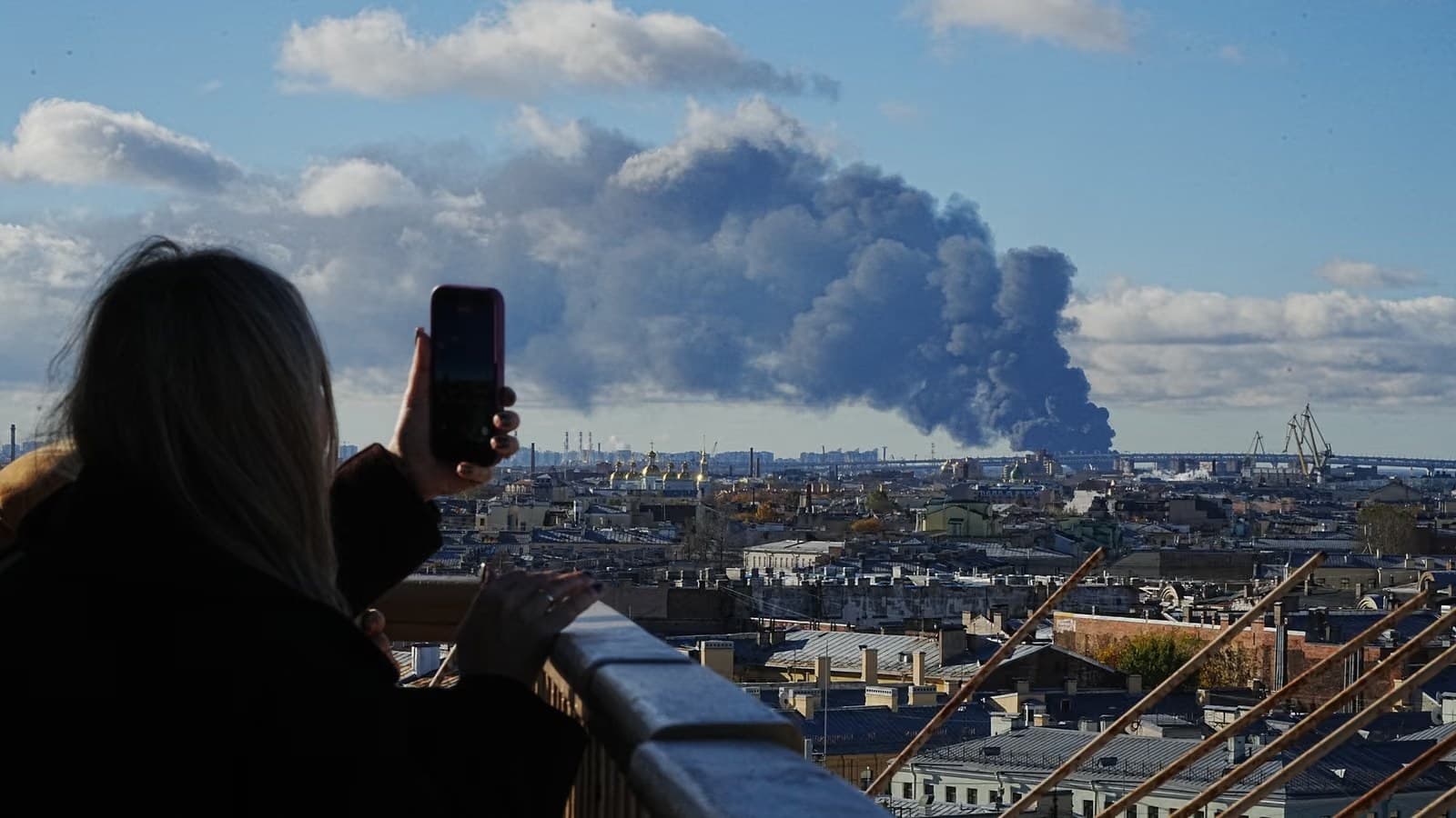 A woman on a rooftop photographs a huge column of grey-black smoke rising over the St. Petersburg skyline.