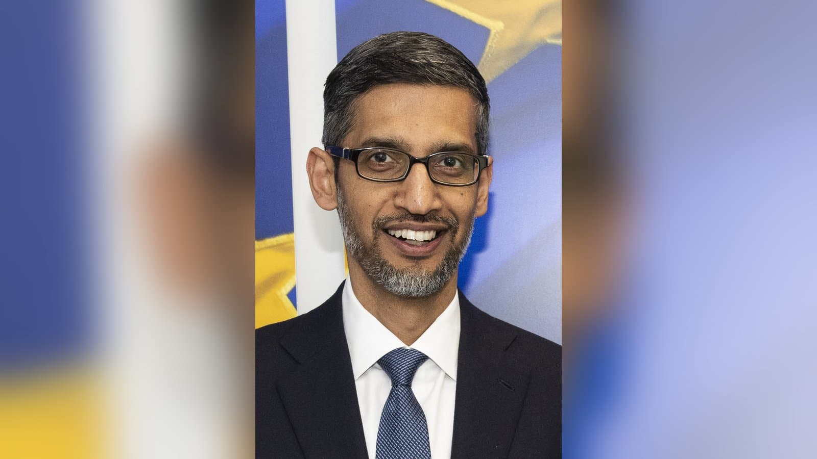 Portrait of Google CEO Sundar Pichai in a dark suit and white shirt, smiling, against a light background.