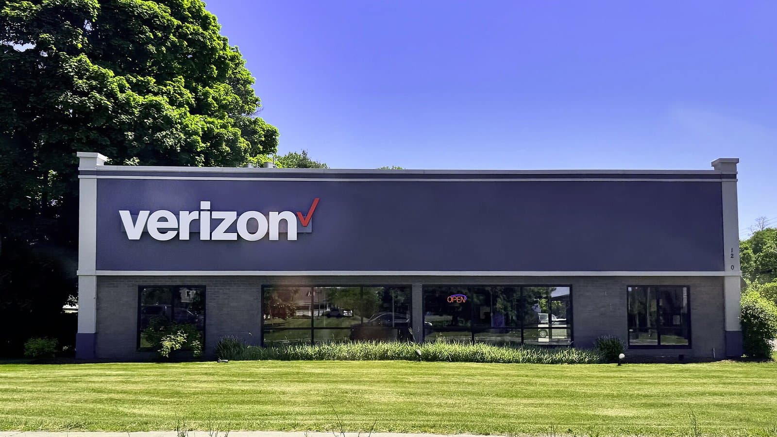 The front of a single-story Verizon retail store with a large white Verizon logo on a dark facade, behind a green lawn under blue sky.
