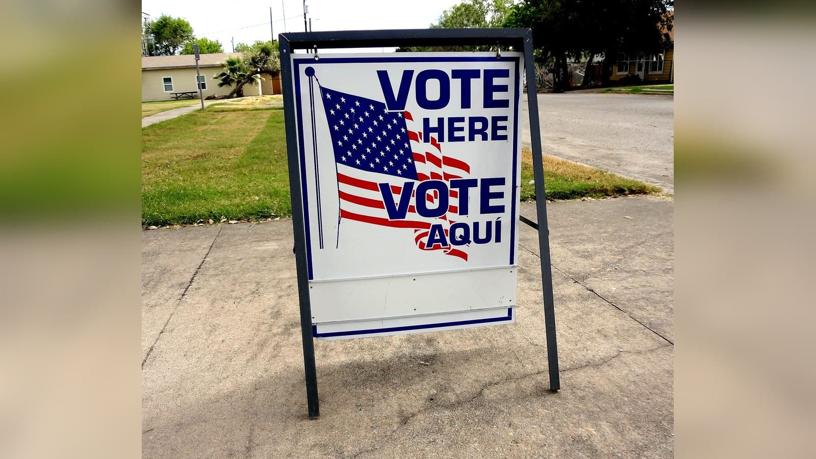 A metal-framed sign reading VOTE HERE and VOTE AQUI with an American flag design, standing on a sidewalk outside a polling site on a sunny day.