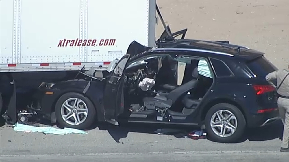 A black Audi SUV with its entire front end sheared away, resting beside the rear of a white semi-trailer on a freeway.